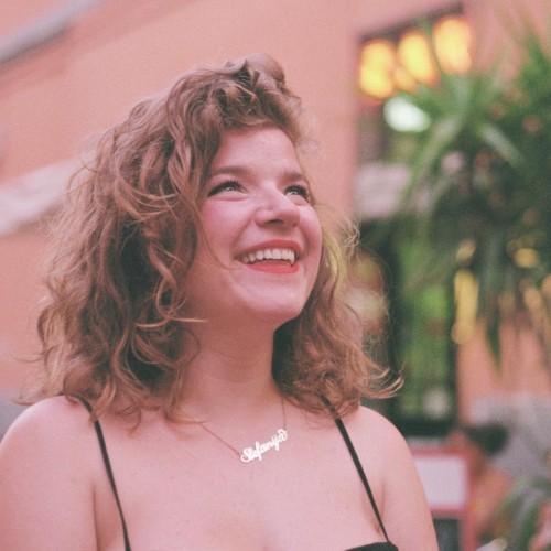 A young, blond woman with curly hair smiling upwards. She is wearing a summer top.