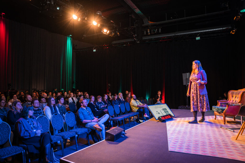 A woman in a berry-coloured dress with a fitting jacket is walking on a stage. People sit in front of the stage, listening to her. You can see the faces of the audience.