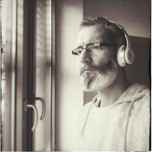 A middle-aged man with glasses, a beard and headphones looks out of a window.